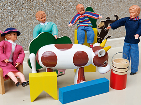 Kleine Menschenpüppchen sitzen auf Miniaturmöbeln. Eine kleine hölzerne Kuh steht im Mittelpunkt.